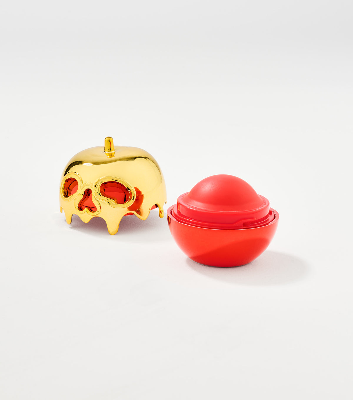 A red spherical lip balm sits open beside its glossy gold dripping skull-shaped apple cap with red eye openings.