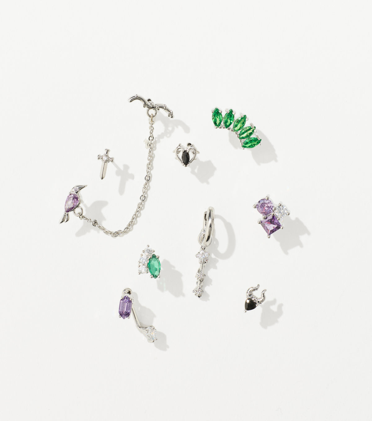 A set of silver earrings with green, purple, and clear gemstones, including a chained bird cuff, dagger stud, and horned heart.