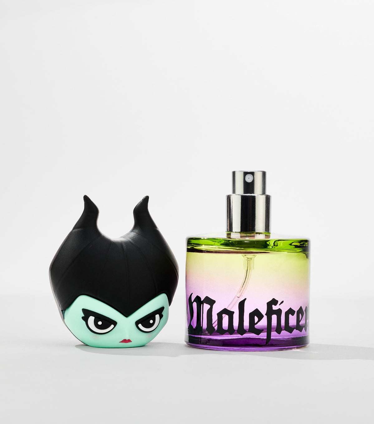 A round glass perfume bottle with a green-to-purple gradient and silver spray nozzle stands beside its horned, green-faced character cap.