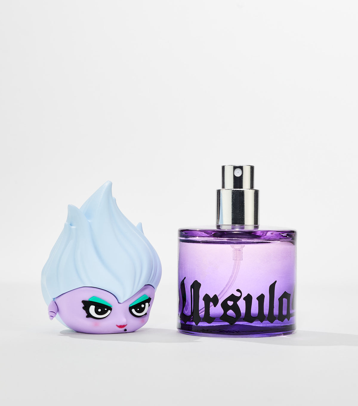 A purple glass perfume bottle with black gothic lettering and silver spray nozzle sits beside its cartoon character cap with pale blue hair.