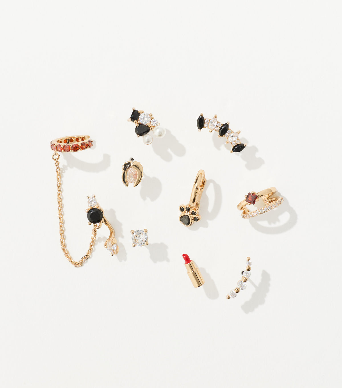 Ten gold-toned earrings with black, red, and clear crystals, including a chain ear cuff, paw hoop, and lipstick stud, are arranged on white.