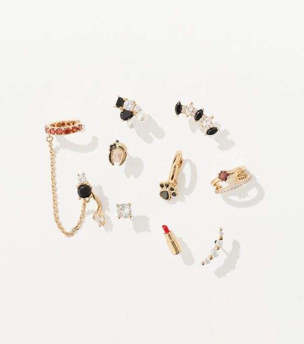 Ten gold-toned earrings with black, red, and clear crystals, including a chain ear cuff, paw hoop, and lipstick stud, are arranged on white.