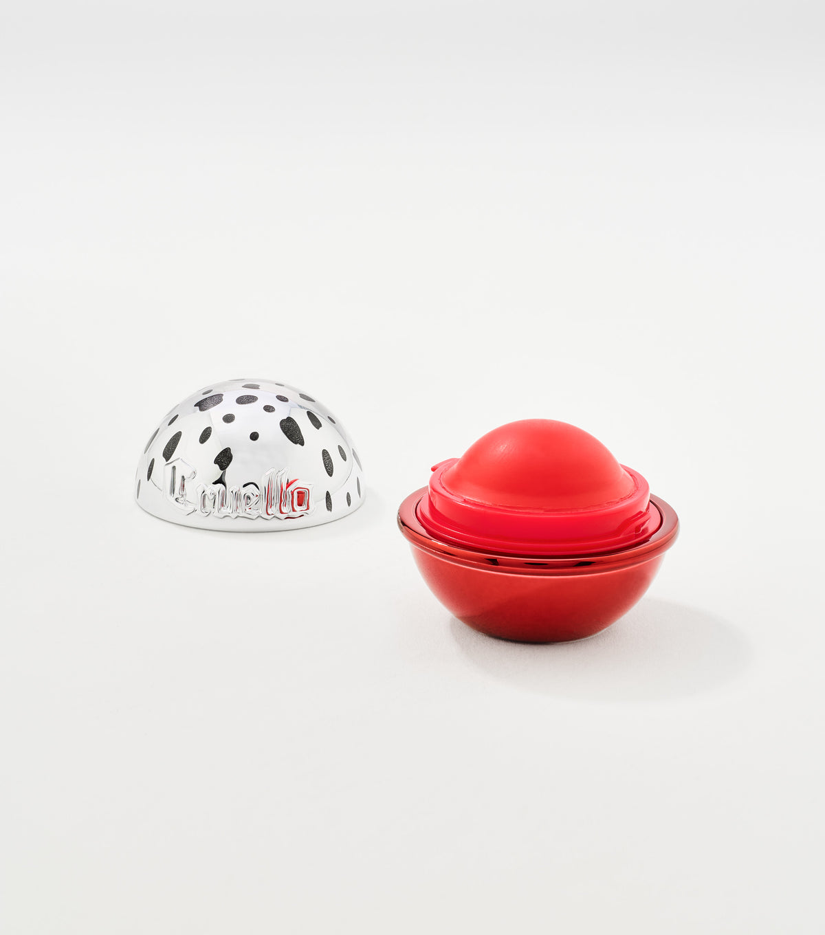 A red spherical lip balm with its glossy red base open beside a silver spotted lid engraved with Cruella.