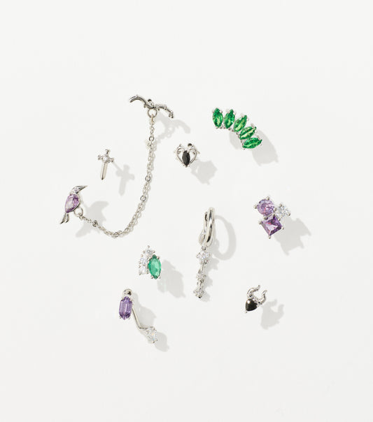 A set of silver earrings with green, purple, and clear gemstones, including a chained bird cuff, dagger stud, and horned heart.