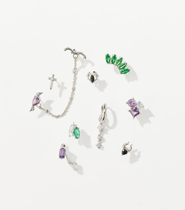 A set of silver earrings with green, purple, and clear gemstones, including a chained bird cuff, dagger stud, and horned heart.