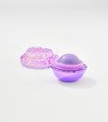 An open spherical lip balm with a purple metallic base and domed balm sits beside its iridescent shell-shaped lid topped with a pearl.