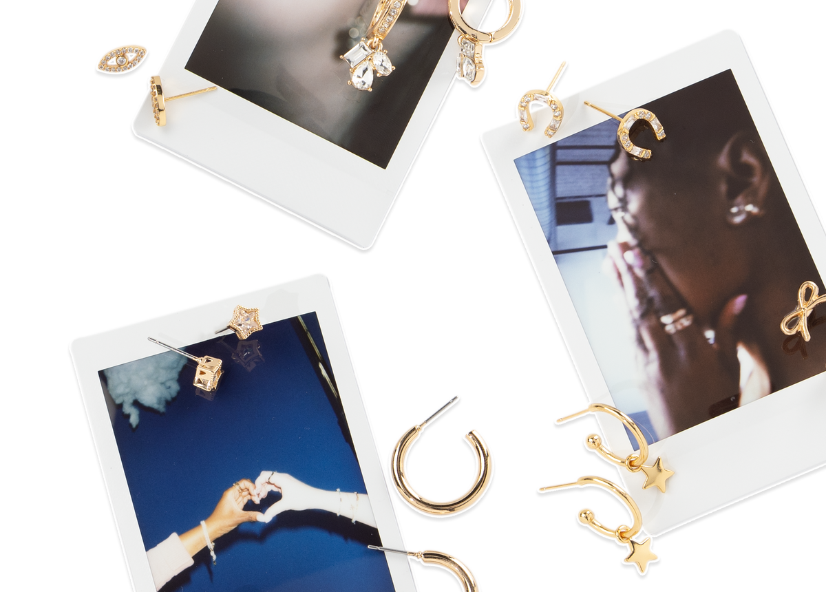 Gold-toned statement earrings and stud earrings displayed around three Polaroid-style photo prints on a white background. The jewelry includes crescent moon, star, and eye-shaped designs, with one earring featuring a clear faceted stone.